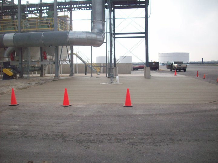 A broom-finished concrete pad at an industrial facility with piping, storage tanks, and a pickup truck in the background.
