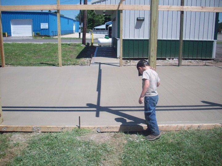 A young boy stands at the edge of a freshly broom-finished plain concrete pad next to a metal building.
