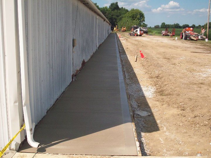 Freshly poured and finished concrete apron running along the base of a large metal agricultural building.