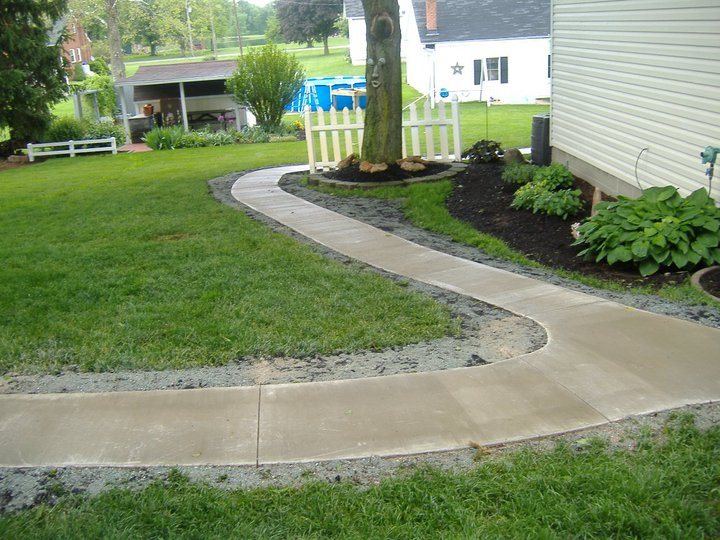 Plain broom-finished concrete walkway curving across a lawn around a tree beside a residential home.