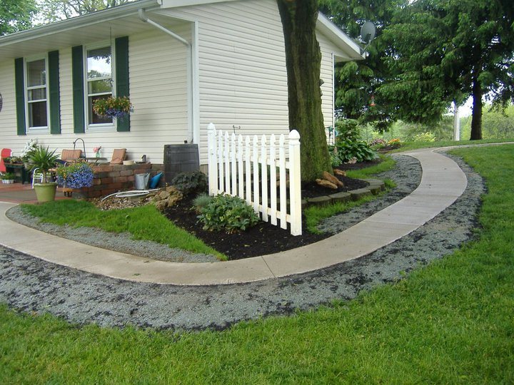 Plain concrete sidewalk curving around the corner of a home past a white picket fence and landscaping.