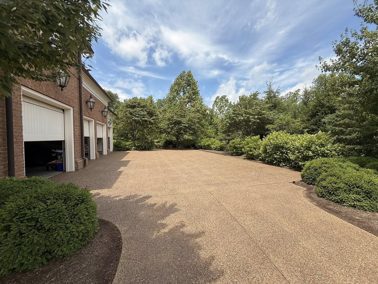 Tan exposed-aggregate concrete driveway beside a brick garage framed by shrubs.