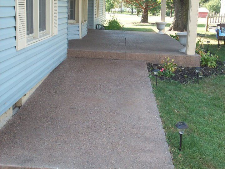 A reddish-brown colored exposed aggregate concrete patio and walkway wrapping around a blue-sided home.