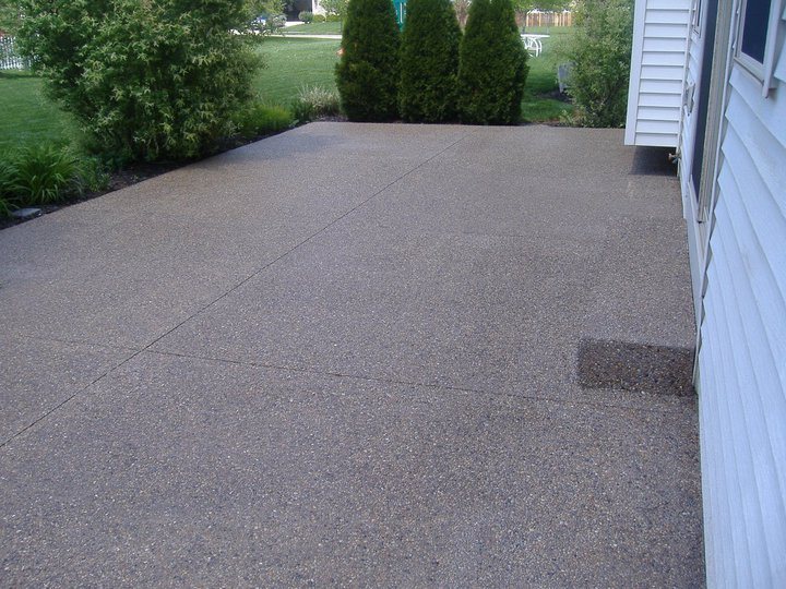 Exposed aggregate concrete patio with visible pebble finish along the side of a vinyl-sided house.