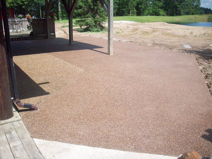 A large finished exposed-aggregate concrete patio under a post-supported roof beside a pond.