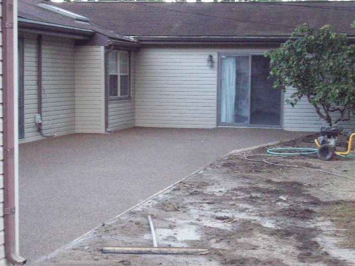 A finished exposed aggregate concrete patio outside a sliding glass door on a sided home, soil being graded.