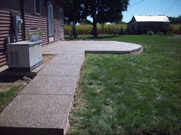 An exposed aggregate concrete patio and connecting walkway beside a rural home with a cornfield and barn behind.