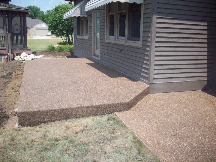 A tan exposed-aggregate concrete patio and adjoining walkway wrap around the corner of a gray-sided home.