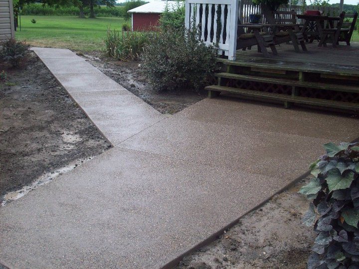 A finished tan exposed-aggregate concrete walkway widens into a patio at the base of a wooden deck.