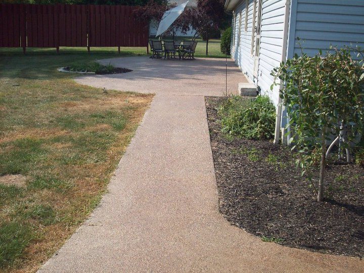 An exposed aggregate concrete walkway leading to a matching patio with a dining set in a fenced backyard.
