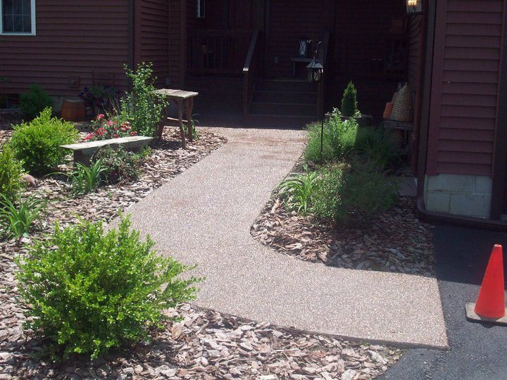 A finished exposed-aggregate concrete walkway leading to the back steps of a red-sided house with landscaping.