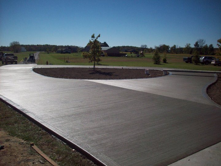 Newly poured broom-finished concrete circular driveway with a center planting island in front of a brick ranch house and detached garage.