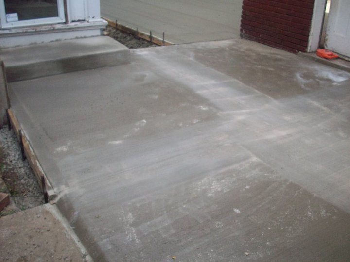 A freshly poured and finished concrete slab with a step against a brick house wall, forms still in place.