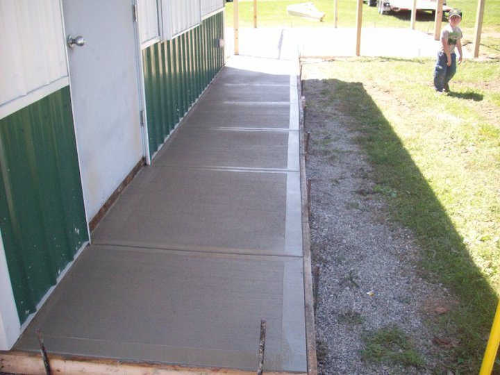 A freshly finished plain concrete sidewalk running alongside a green and white metal building.