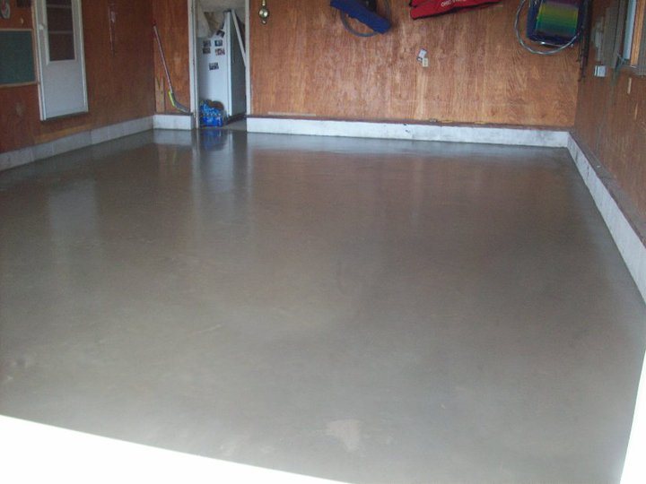 A freshly troweled smooth concrete garage floor inside a wood-paneled garage, photographed from the opposite corner.