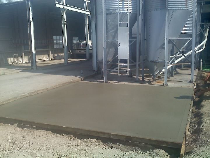 A freshly poured smooth concrete equipment pad beneath grain bins and feed silos at an agricultural facility.