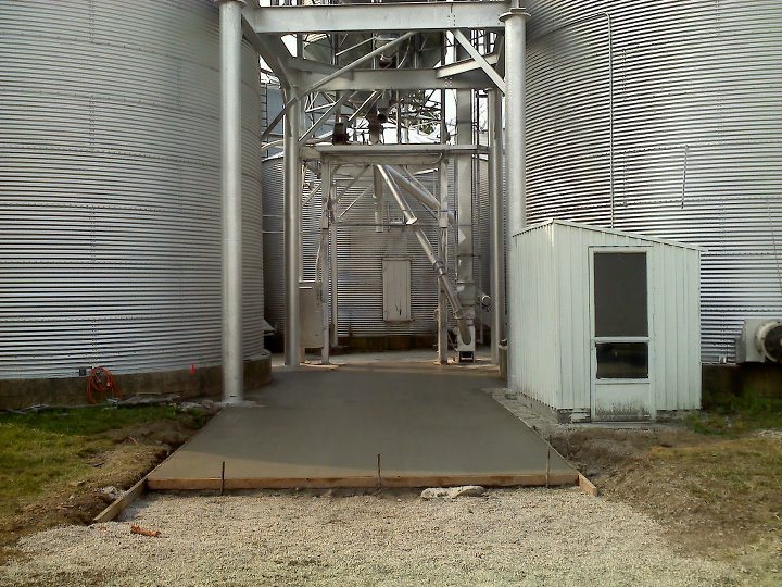 A freshly poured concrete walkway pad between large galvanized grain bins at a farm grain facility.