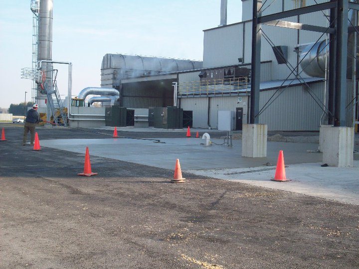 A freshly poured concrete pad at an industrial processing plant marked with orange traffic cones and a worker nearby.