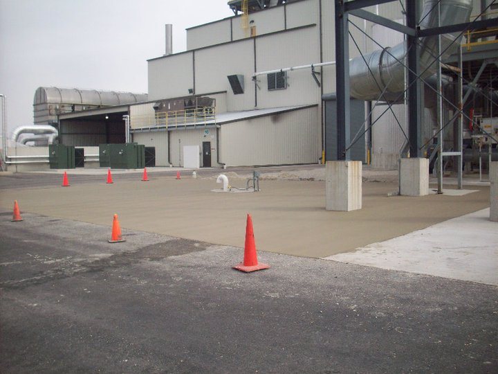 A large fresh concrete pad poured beside a metal industrial plant building on an overcast day with cones set out.
