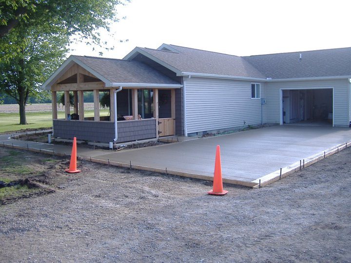 Large freshly finished concrete patio slab in forms behind a home with a covered porch, marked with cones.