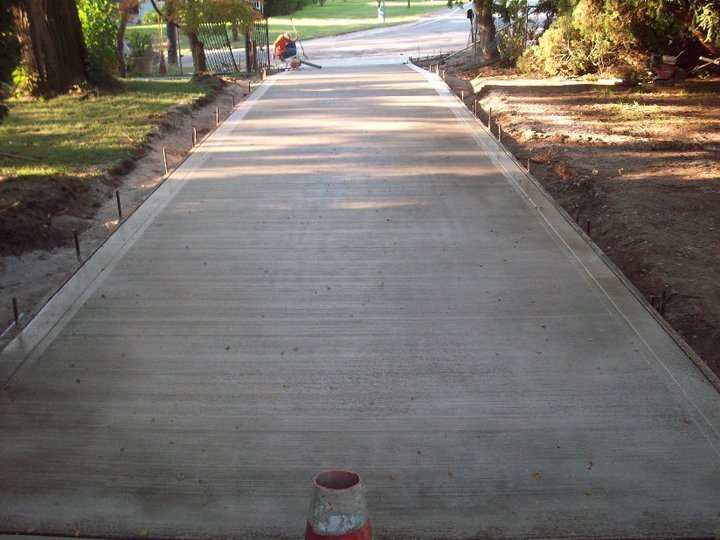 A long freshly finished plain concrete driveway with broom-finish texture and rebar pins along both edges.