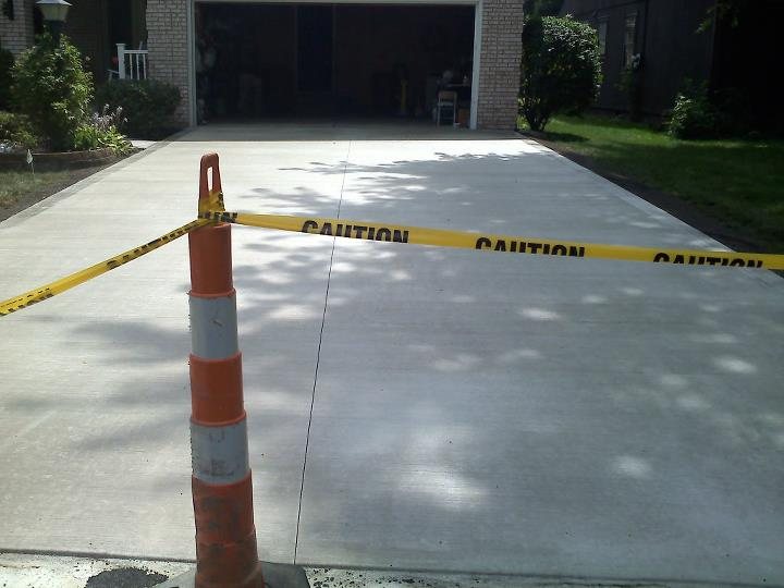 A freshly finished plain gray concrete driveway taped off with caution tape leading to a brick home's garage.