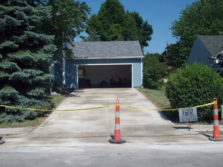 Newly poured plain concrete driveway leading to a blue detached garage, taped off with a Custom Crete yard sign.