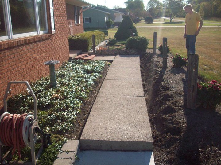 Finished plain gray concrete sidewalk running alongside a brick house with flower beds and a man standing nearby.