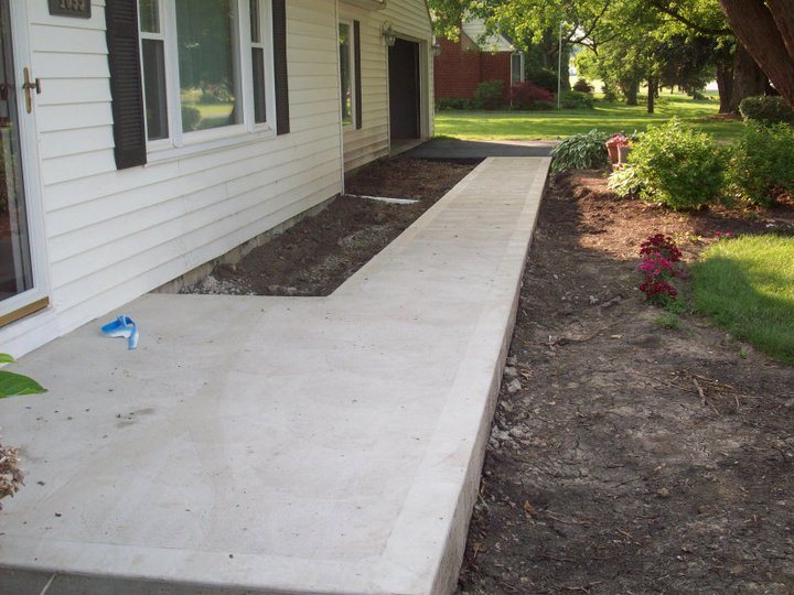 Newly poured plain gray concrete walkway running alongside a white sided house with fresh landscaping.