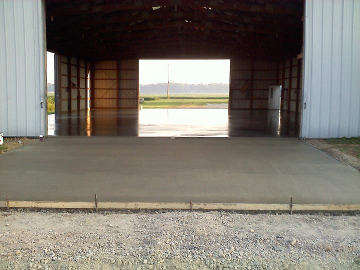 A freshly finished concrete floor poured inside a rural pole barn open at both ends to farm fields.