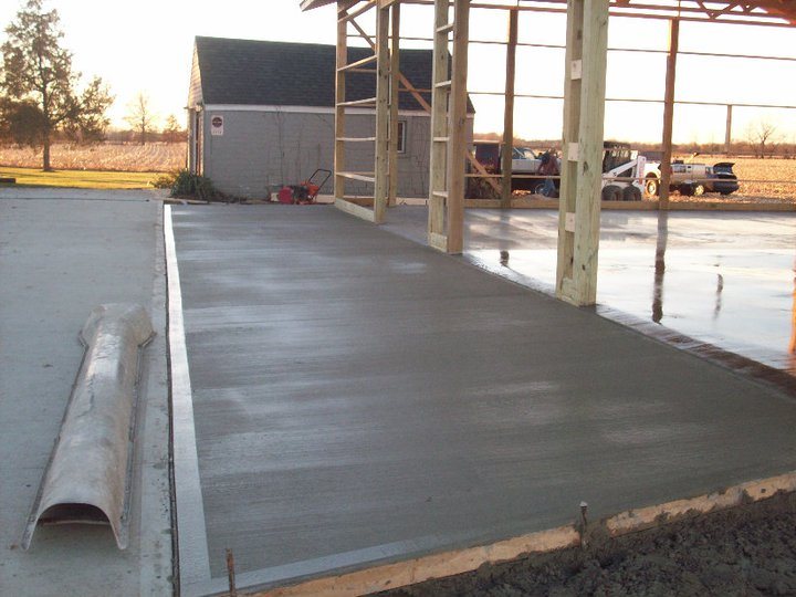 A freshly broom-finished concrete slab poured under a wood-framed pole barn at sunset with trucks parked beyond.