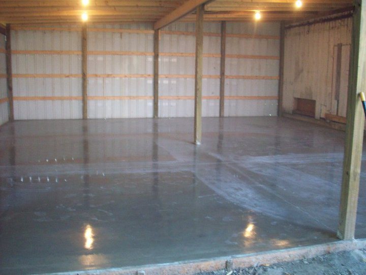 Newly finished concrete slab with a wet sheen curing inside a lit metal pole barn.