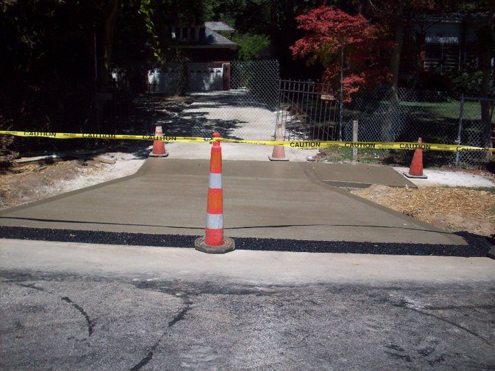 A newly poured plain concrete driveway apron meeting the street, blocked off with caution tape and orange cones.