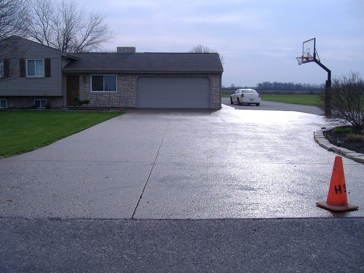 A finished plain concrete residential driveway leading to a two-car garage, with a basketball hoop and traffic cone.