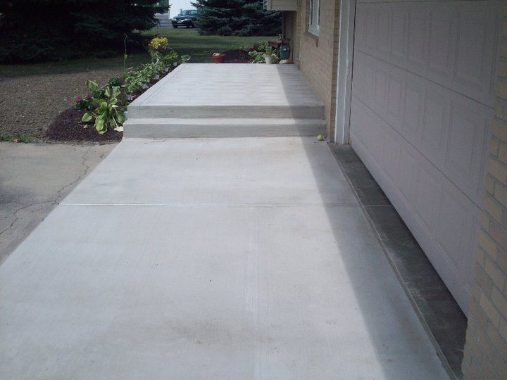 Finished plain concrete driveway leading to a raised entry landing beside a tan brick house garage.