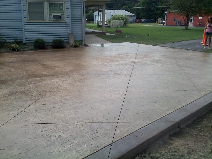 A tan seamless-textured stained concrete patio with a darker brown stamped border beside a blue house.