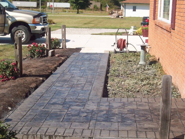 Charcoal-gray stamped concrete walkway with an ashlar pattern and brick border running alongside a brick house with a truck parked nearby.
