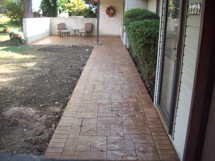 Tan stamped concrete walkway with an ashlar-slate pattern and brick-stamped border leading along a house with a matching stamped patio.