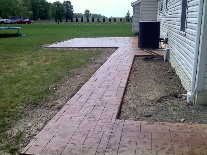 Red running-bond brick-pattern stamped concrete walkway leading to a stamped patio along a house.