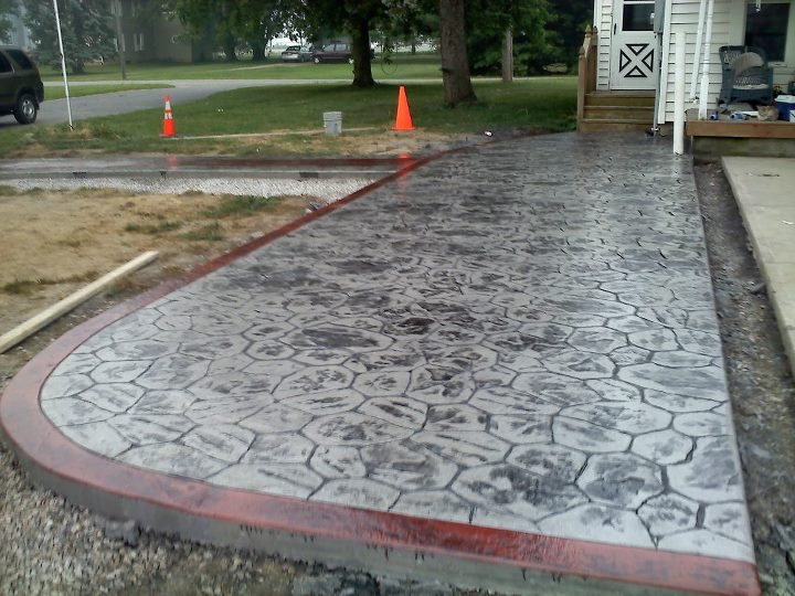 A gray cobblestone-pattern stamped concrete driveway with a red-stained border and safety cones in front of a house.