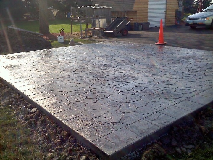 Dark gray random-flagstone stamped concrete patio slab with a soldier-course border and an orange cone on top, soil not yet backfilled.