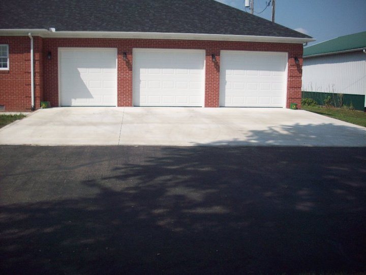 A finished plain concrete driveway apron in front of a three-bay brick garage transitioning to a blacktop drive.