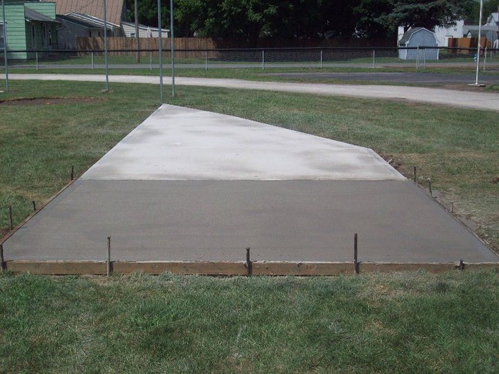 Freshly finished trapezoidal concrete pad still in forms in a grassy area near a residential street.