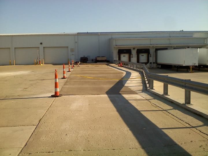 A concrete loading-dock approach and drive lane at a commercial warehouse with overhead doors, dock trailers, and guardrail.