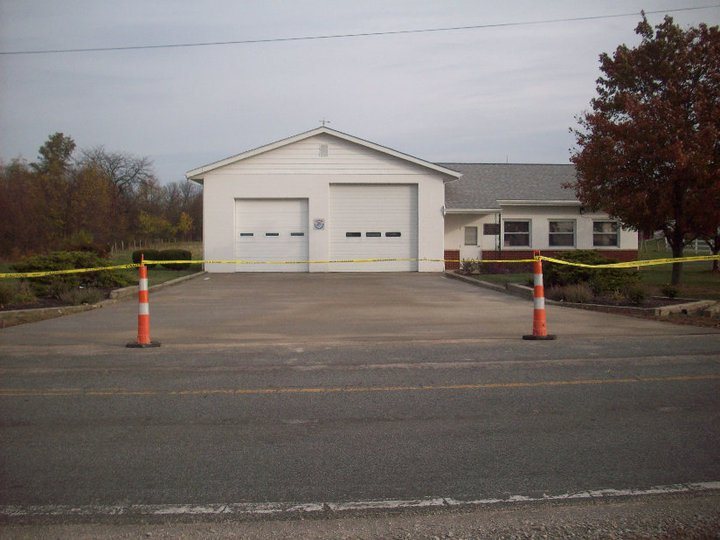 A white two-bay garage with fresh concrete apron cordoned off by traffic cones and yellow caution tape while curing.