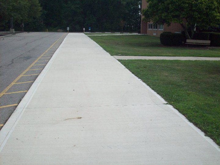 Long wide plain concrete sidewalk running alongside a parking lot beside a brick commercial building.