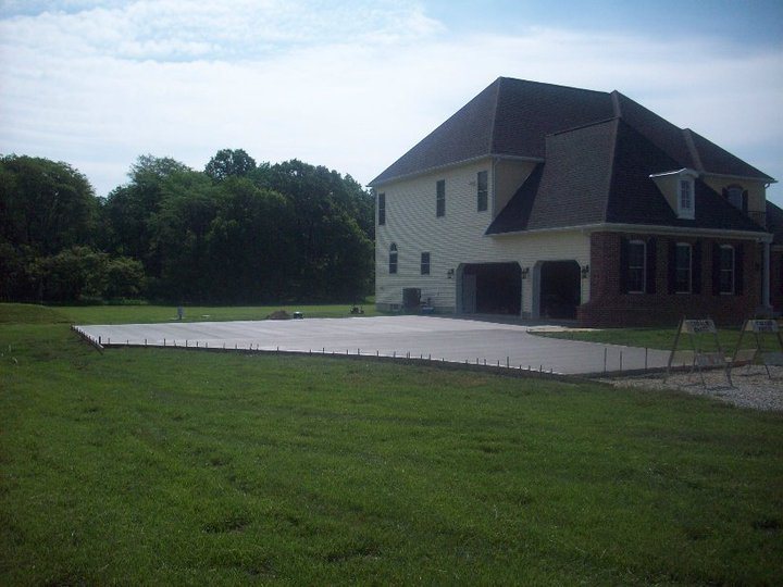 A wide finished plain concrete driveway apron at a large rural home, shown from across the lawn.