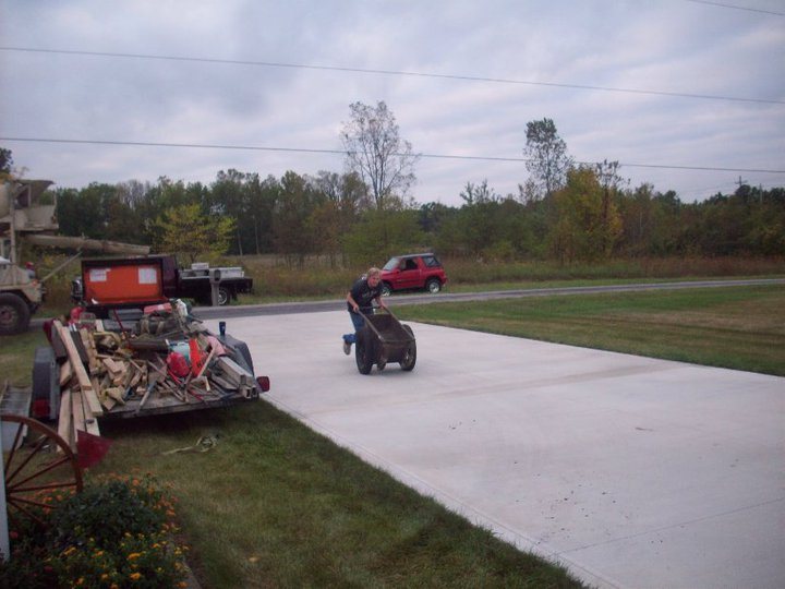 Finished plain gray concrete driveway in a rural setting with a man pushing a wheelbarrow and an equipment trailer parked at the edge.