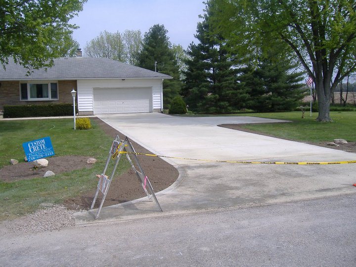 Cured plain concrete residential driveway with a parking apron beside a ranch home, roped off with caution tape and a Custom Crete sign.